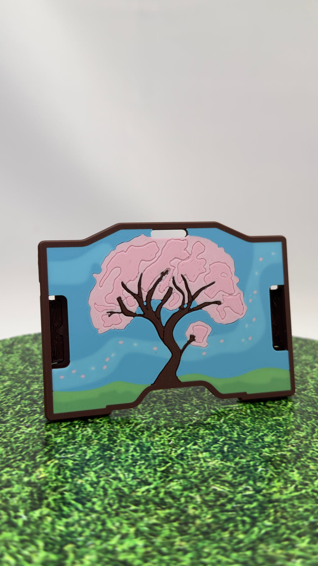 A keycard holder featuring a printed cherry blossom tree design with pink blossoms, green hills, and a blue sky, shown standing on a surface with a blurred background.