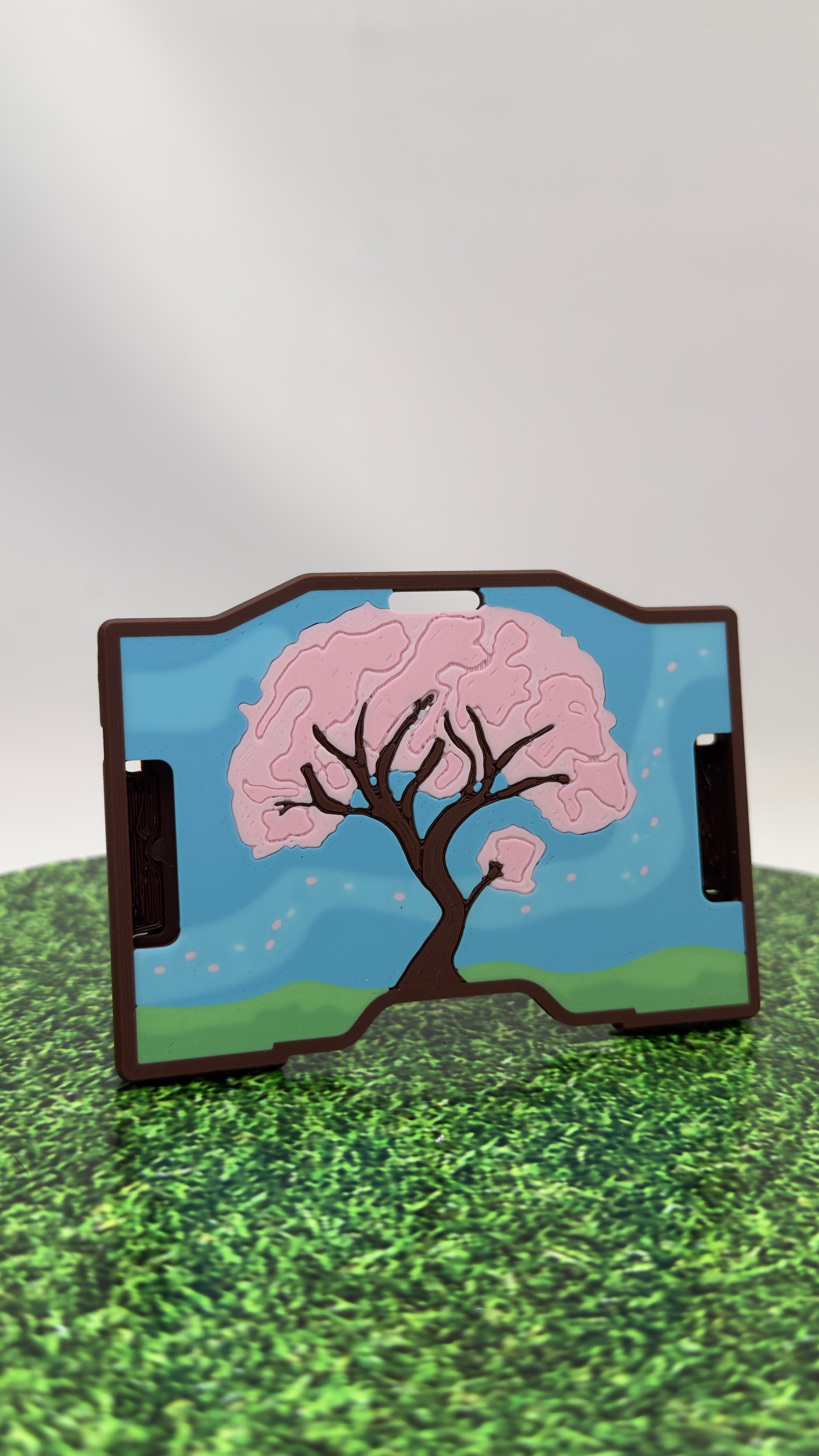 A keycard holder featuring a printed cherry blossom tree design with pink blossoms, green hills, and a blue sky, shown standing on a surface with a blurred background.