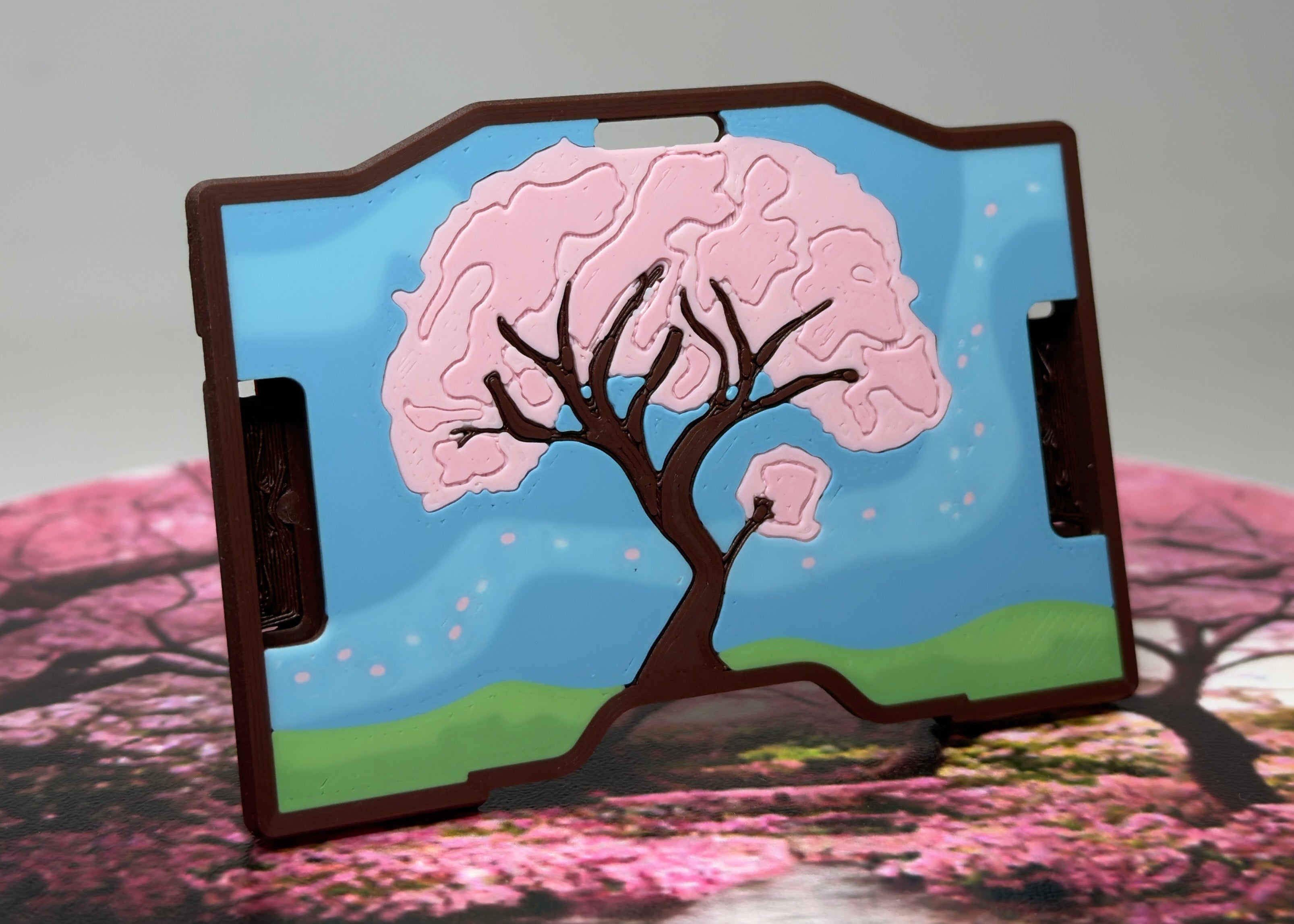 A keycard holder featuring a printed cherry blossom tree design with pink blossoms, green hills, and a blue sky, shown standing on a surface with a blurred background.