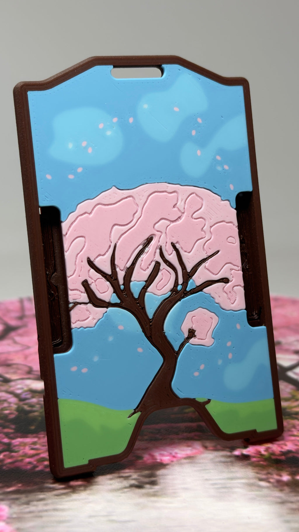 A keycard holder with a printed cherry blossom tree design, standing on a pink and white surface against a light gray background.
